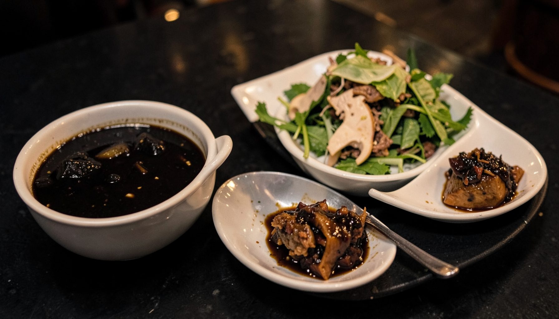 A spread of Surabaya street dishes on a dark surface: a bowl of black Rawon soup, a plate of Rujak Cingur with sliced snacks