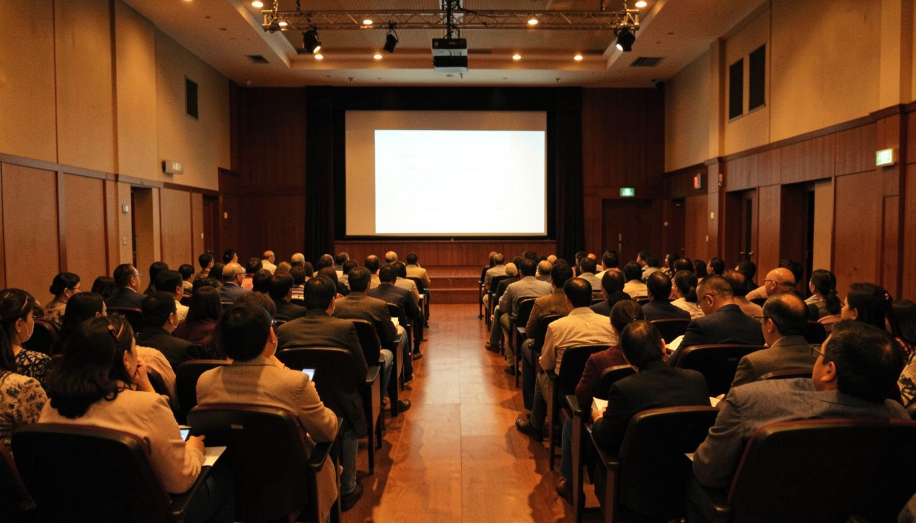 A warmly lit seminar hall in Surabaya with rows of seated attendees facing a presentation stage, bathed in amber and deep brown tones