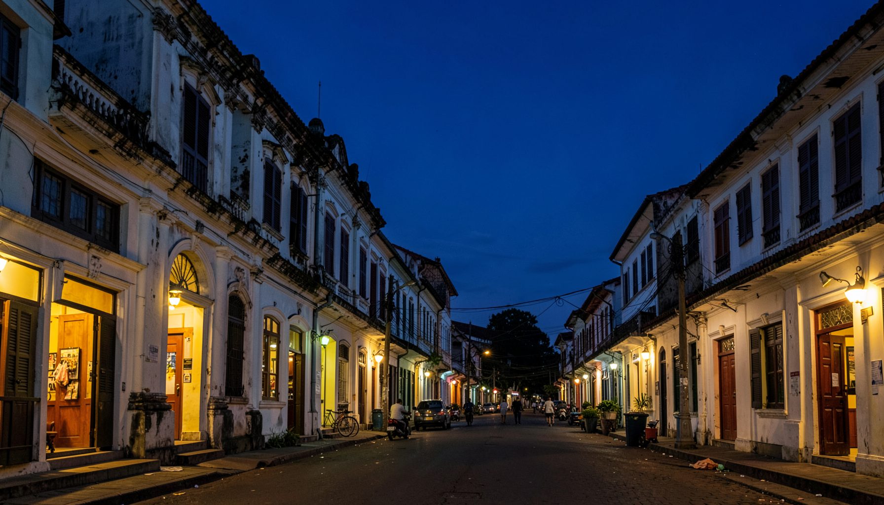 A wide atmospheric shot of Surabaya's old-town streets at dusk, colonial-era buildings with warm yellow light spilling from doorways