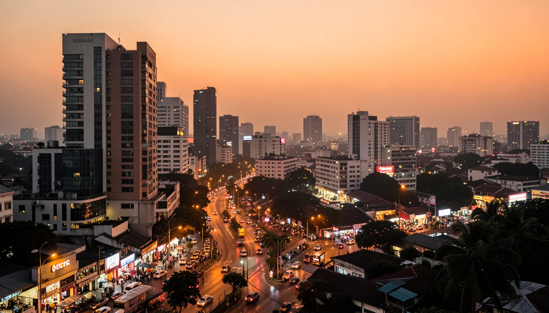 A wide cityscape of Surabaya at dusk, with warm amber streetlights reflecting off modern buildings and a hazy orange sky